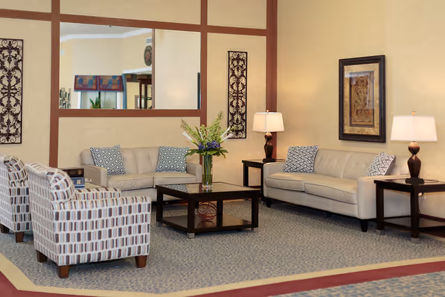 A cozy living room area with two beige sofas and two patterned armchairs arranged around a dark wooden coffee table with a vase of flowers. The room features beige walls with decorative wall art, two side tables each with a lamp, and a large mirror on the wall reflecting part of the room.