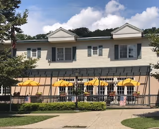 Exterior view of a two-story senior living facility building with beige siding and green shutters. The ground floor features a patio area with several yellow umbrellas and outdoor seating. There are small American flags displayed along the patio railing, and a tree is visible on the left side. The sky is partly cloudy.