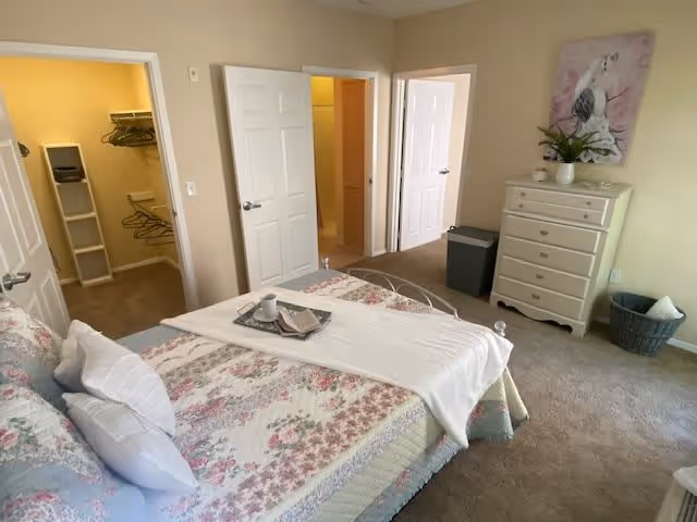 A bedroom with a floral patterned bedspread on a bed, white pillows, and a white runner. There is a tray with a cup and napkin on the bed. The room has beige walls and carpet. To the left, there is an open door leading to a walk-in closet with hangers and shelving. In the center, another open door leads to a bathroom. On the right side, there is a white dresser with a plant and decorative items on top, a trash bin, and a laundry basket. A framed picture of a bird hangs above the dresser.