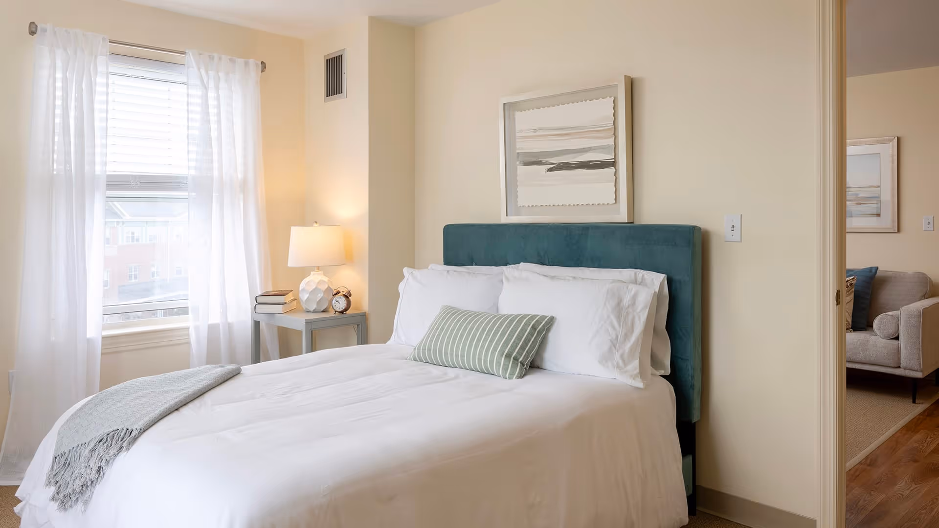 A bright bedroom with a neatly made bed featuring white linens, a green upholstered headboard, and a green striped pillow. A small bedside table with a lamp, books, and an alarm clock is next to the bed. Sheer white curtains cover the window, allowing natural light to fill the room. A doorway reveals a glimpse of a living room with a beige sofa and framed artwork on the walls.