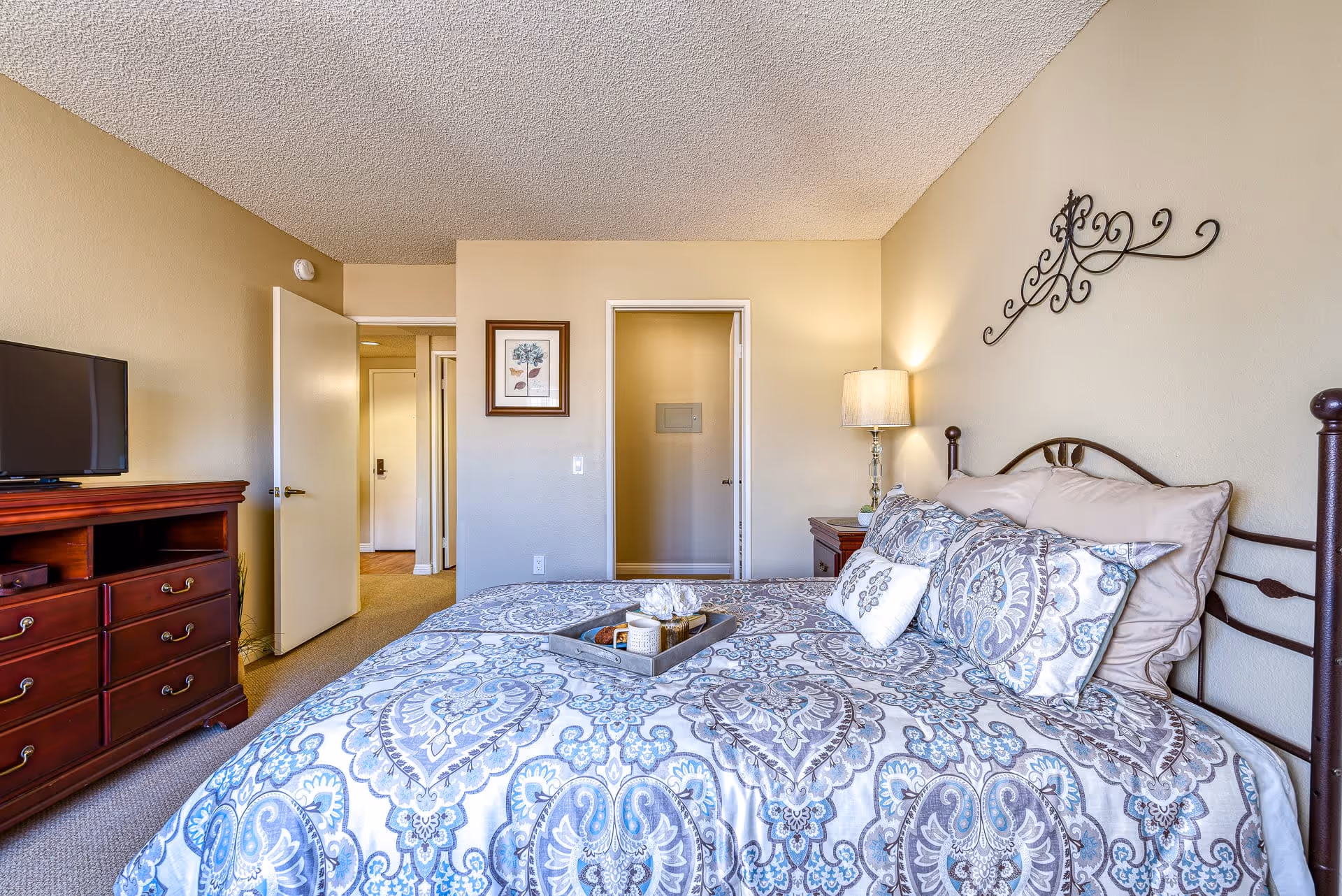 A bedroom in a senior living facility with a bed covered in a blue and gray patterned comforter and multiple pillows. There is a wooden dresser with a TV on top to the left, a nightstand with a lamp to the right, and a decorative metal wall hanging above the bed. The room has beige walls and carpeted floor, with an open door leading to a hallway and another door leading to a closet or bathroom.