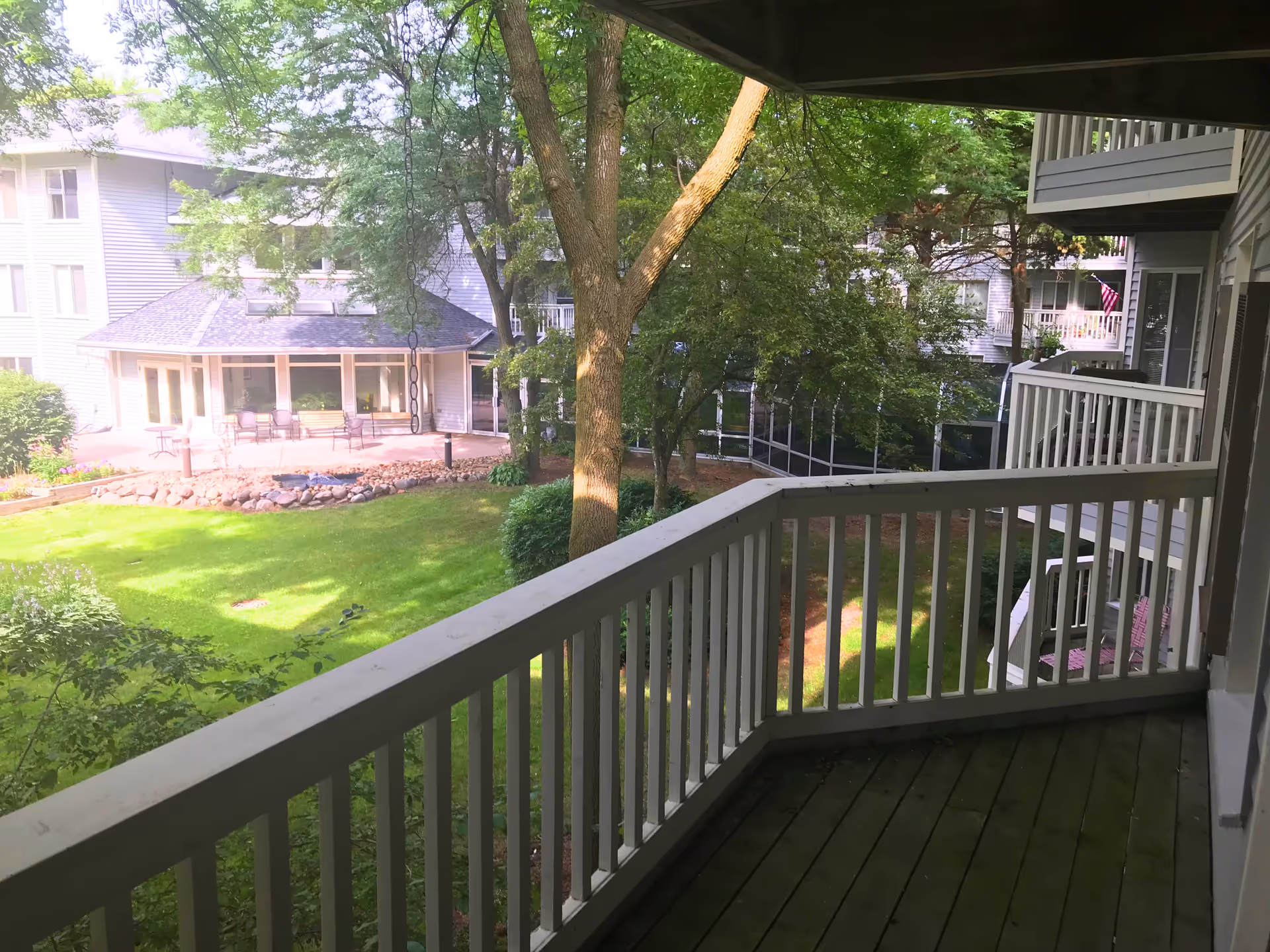View from a balcony overlooking a green courtyard with trees and grass. Surrounding the courtyard are multi-story buildings with balconies and a patio area with chairs and tables.
