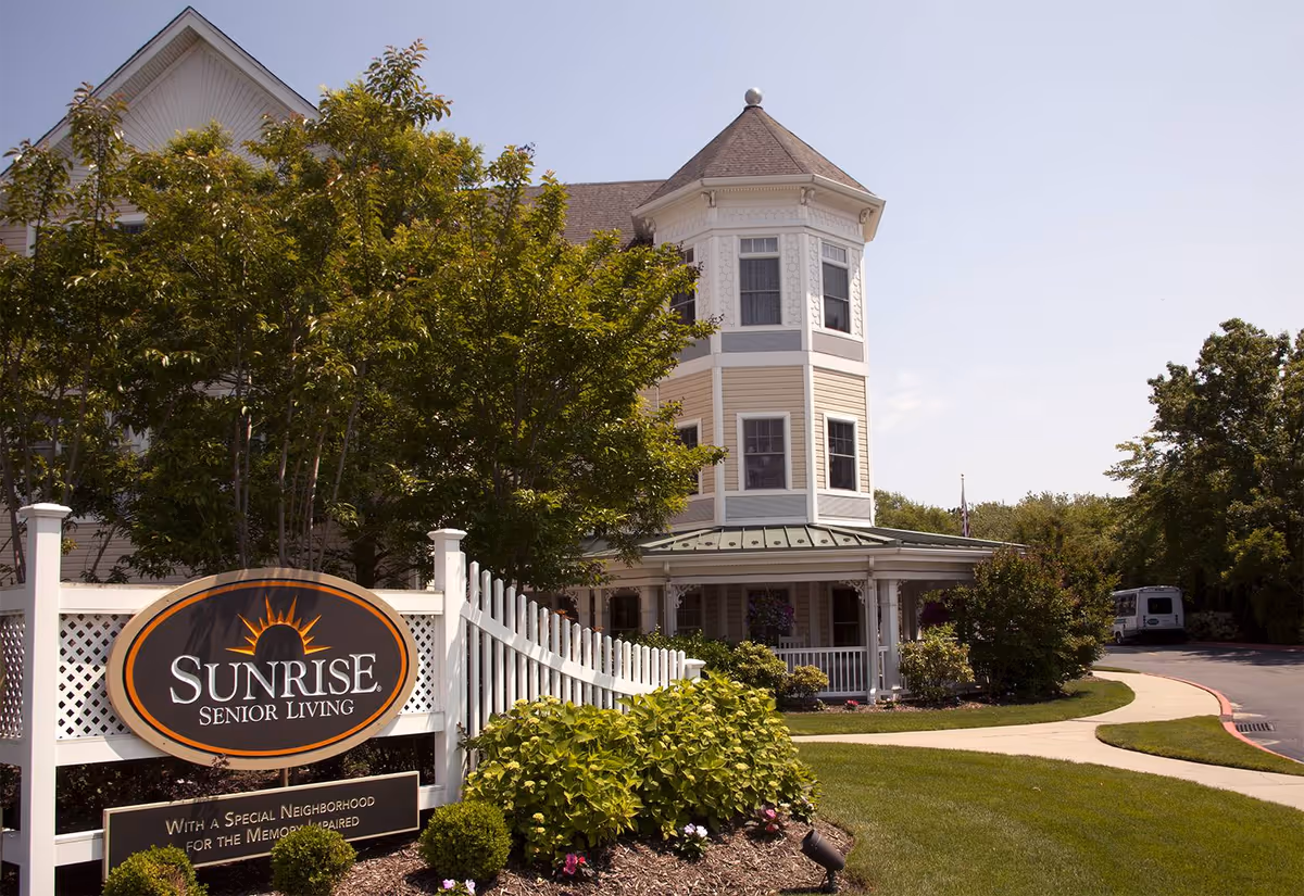 Exterior view of Sunrise Senior Living facility showing a large, light-colored building with a turret and wraparound porch, surrounded by trees and landscaped greenery. A white picket fence with a Sunrise Senior Living sign is visible in the foreground.