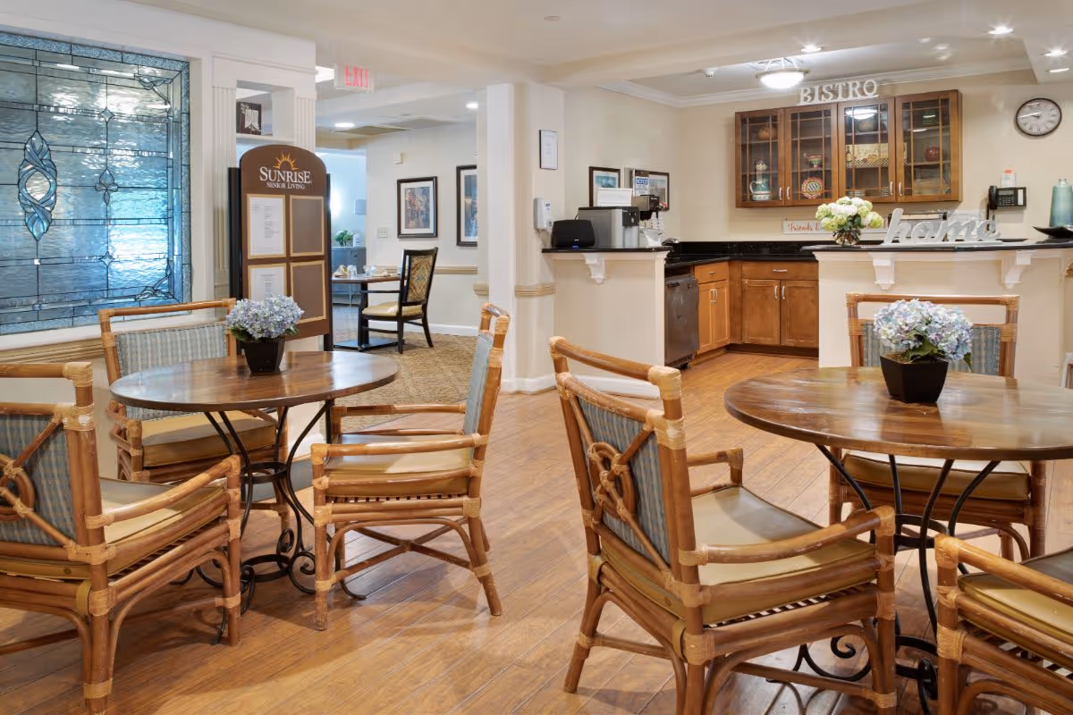 A cozy dining area in a senior living facility with round wooden tables and cushioned chairs. Each table has a small potted flower arrangement. In the background, there is a kitchen counter with wooden cabinets and a sign that reads 'BISTRO'. A decorative stained glass window and a Sunrise Senior Living sign are visible on the left side of the image.