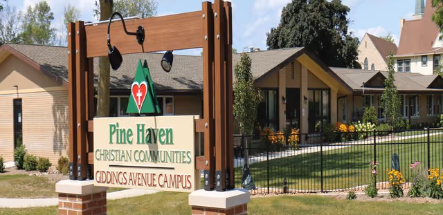Outdoor view of Pine Haven Christian Communities Giddings Avenue Campus sign in front of a single-story building with a lawn, flowers, and trees under a blue sky with some clouds.