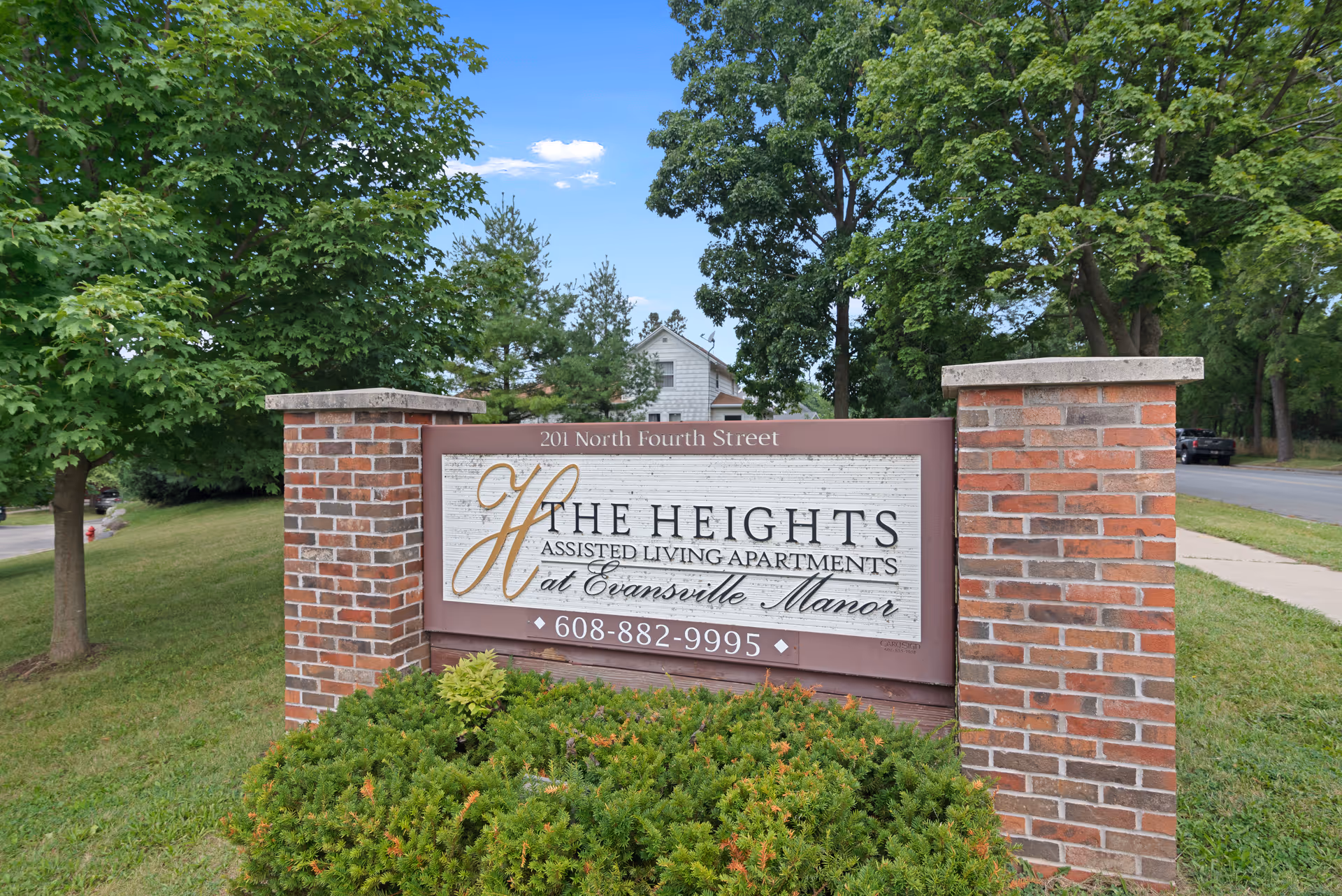 A brick sign for The Heights Assisted Living Apartments at Evansville Manor located at 201 North Fourth Street, surrounded by green bushes and trees with a clear blue sky in the background.