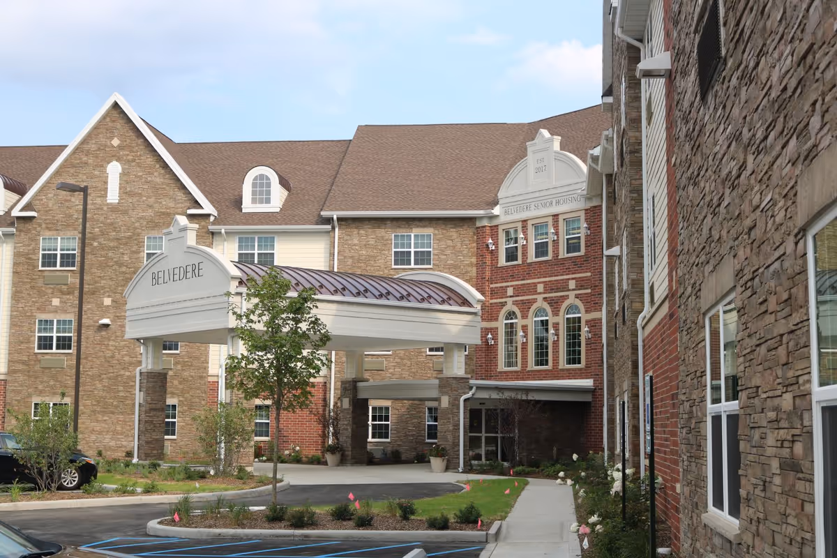 Exterior view of Belvedere Senior Housing building showing a covered entrance with the name Belvedere on it, multiple windows, and a landscaped area with small trees and bushes in front.