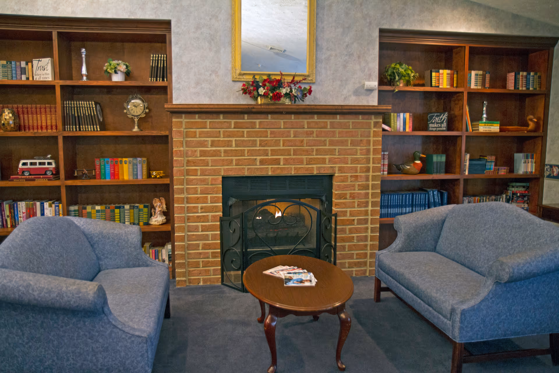 Cozy sitting area with two blue upholstered armchairs facing each other across a wooden coffee table with magazines on it. Behind the table is a brick fireplace with a fireguard and a floral arrangement on the mantel. Flanking the fireplace are built-in wooden bookshelves filled with books, decorative items, and plants. A large mirror hangs above the fireplace.