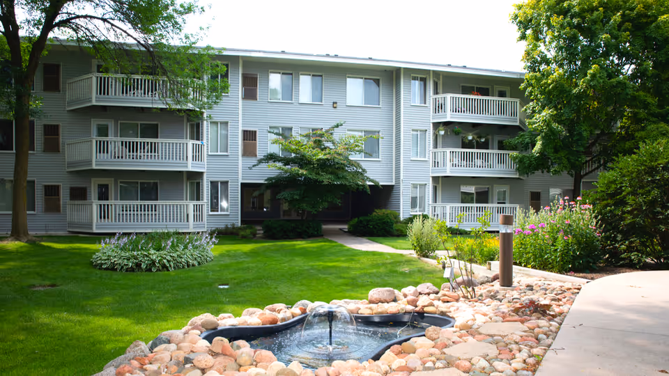 Exterior view of a senior living facility building with multiple balconies, surrounded by green grass, trees, flowering plants, and a small decorative pond with a fountain in the foreground.