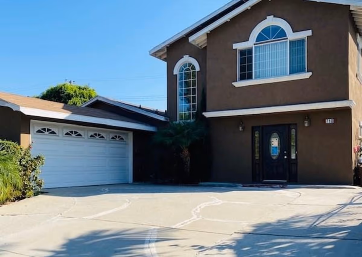 Exterior view of a two-story brown house with white trim, featuring a large driveway, a double garage door, and arched windows above the front door and on the second floor.