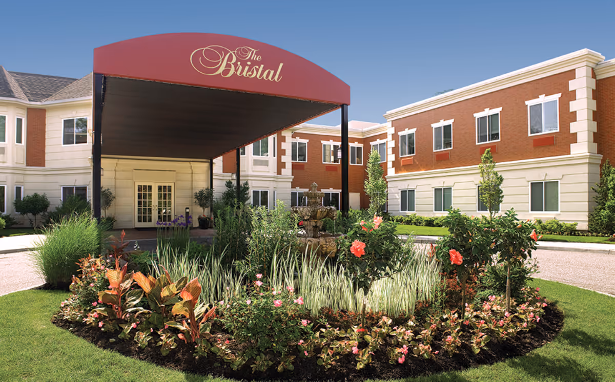 Front exterior view of The Bristal Assisted Living at Massapequa building with a red canopy entrance, landscaped garden with flowers and shrubs, and a multi-tiered fountain in the center.