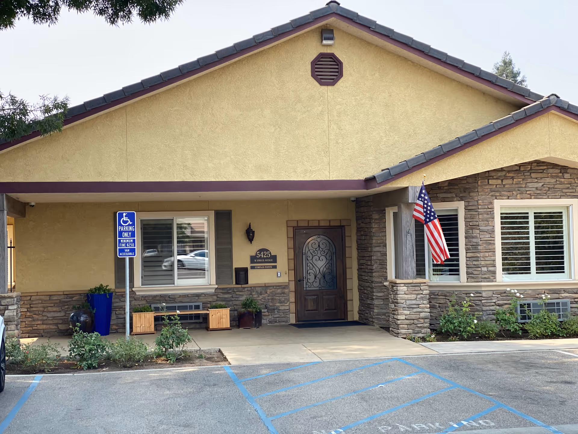 Front exterior view of a single-story building with beige stucco walls and stone accents. There is a wooden door with decorative glass, two windows with white shutters, an American flag mounted on a stone column, and a blue handicapped parking sign in front. The building has a sloped roof with dark shingles.