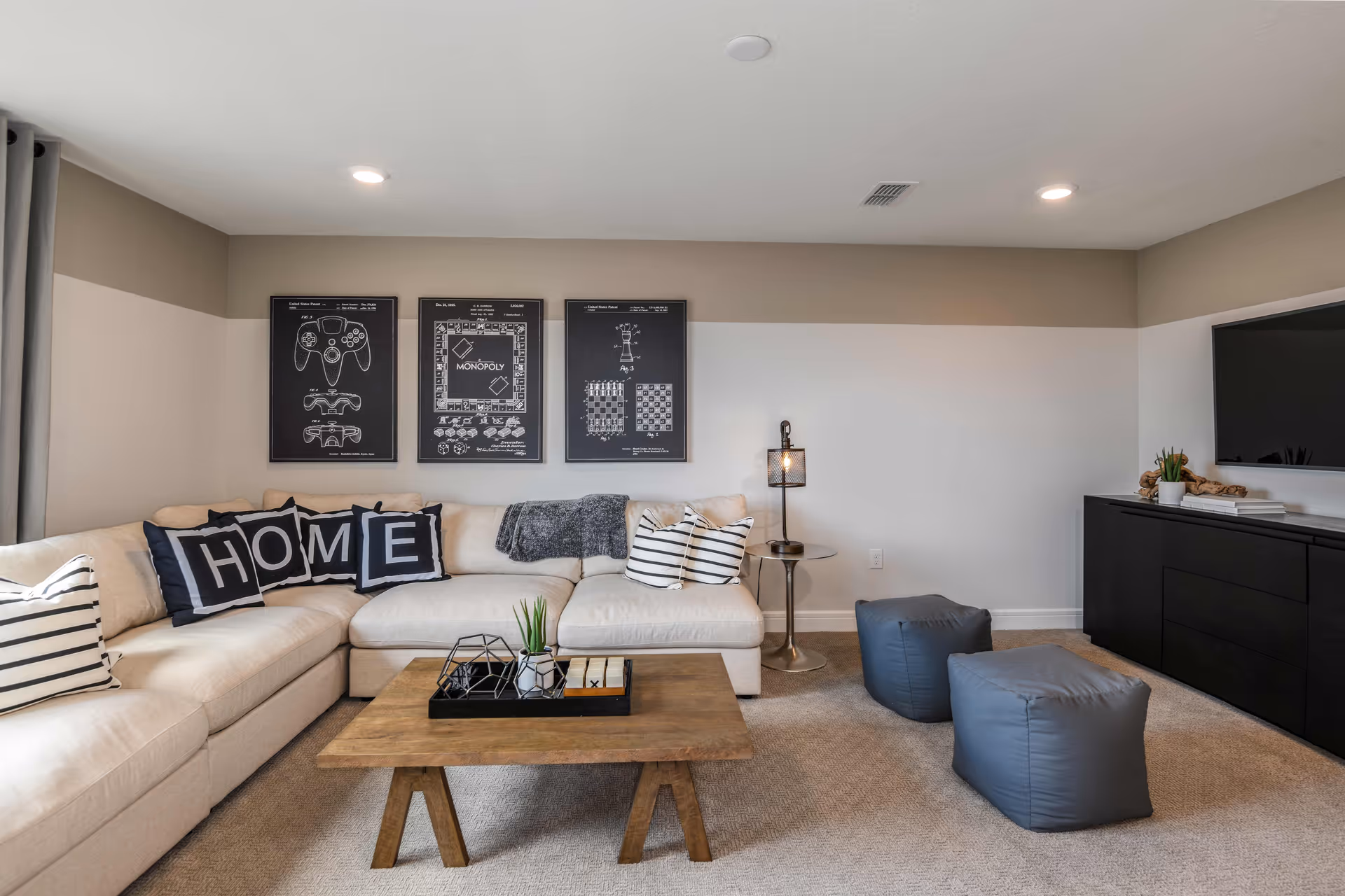 A cozy living room with a beige sectional sofa adorned with pillows, including four black pillows spelling out HOME. Above the sofa are three framed black and white prints featuring game-related designs. A wooden coffee table with decorative items sits in front of the sofa. To the right, there are two gray poufs on a carpeted floor, a small round side table with a lamp, and a black media console with a flat-screen TV mounted on the wall above it. The walls are painted in two tones of beige and white.