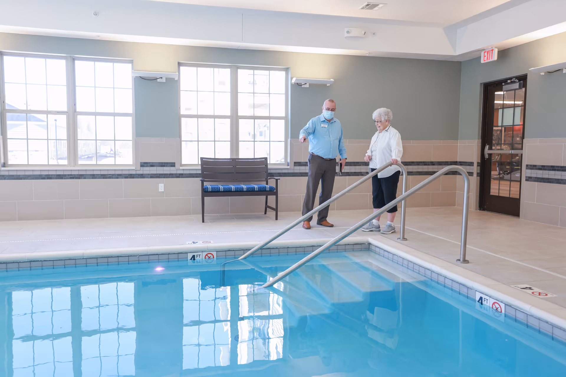 An indoor swimming pool area with a man wearing a mask and an elderly woman standing near the pool's edge. The pool has metal handrails and steps leading into the water. There is a bench with a blue cushion against the wall, large windows letting in natural light, and a door with an exit sign above it.