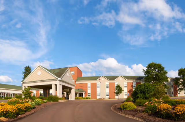 Exterior view of a senior living facility named Suites At Rouse, featuring a large driveway leading to a covered entrance. The building is a multi-story structure with a combination of brick and light-colored siding. The surrounding landscape includes well-maintained flower beds and shrubs under a partly cloudy blue sky.