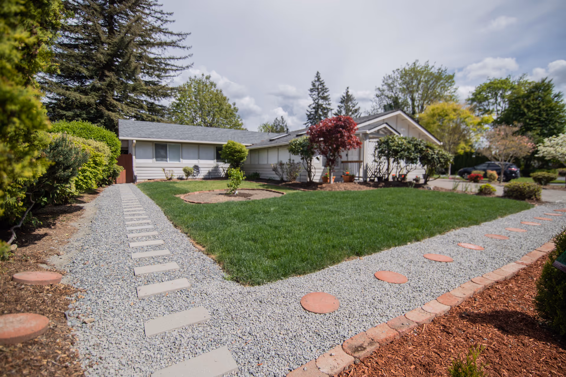 Exterior view of a single-story residential building with a well-maintained lawn and landscaped garden. There are two gravel pathways with stepping stones leading to the entrance of the building. Various trees and shrubs surround the property under a partly cloudy sky.