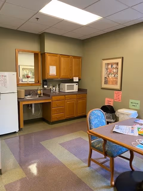 A small kitchen area in a retirement community with wooden cabinets, a microwave, a sink, and a white refrigerator. There is a table with chairs, newspapers, and a face mask on it. The walls are light green with a framed floral picture and signs indicating trash and recycling bins.