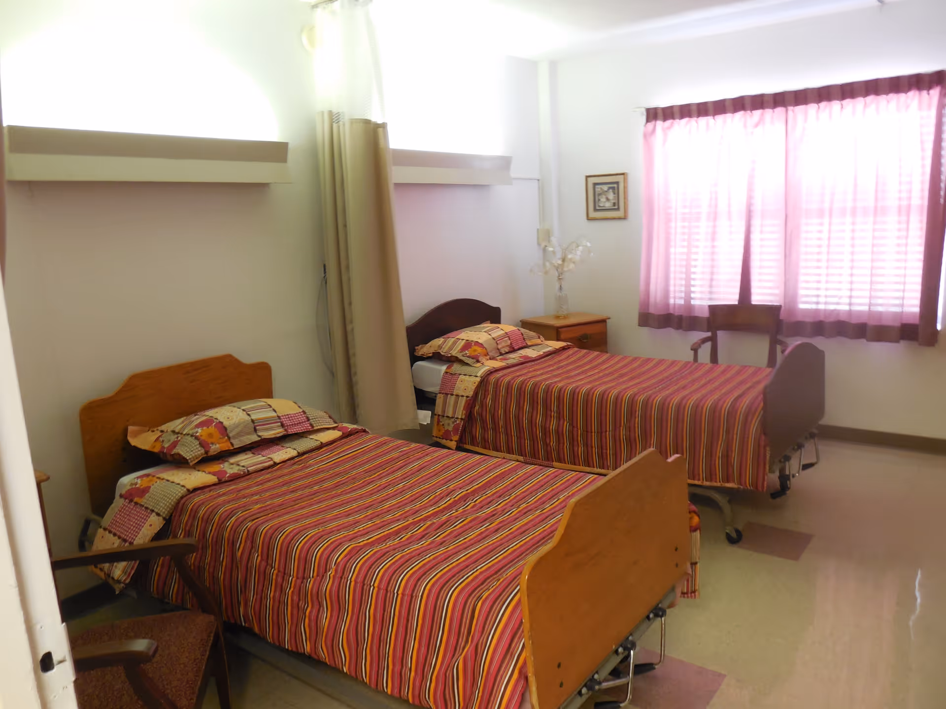 A nursing center bedroom with two single beds, each with striped bedspreads and matching pillows. There is a wooden chair near the foot of one bed and a nightstand with a decorative plant between the beds. A window with pink curtains allows natural light into the room.