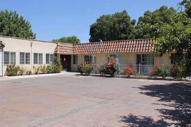 Exterior view of a single-story building with beige walls and a red tile roof, surrounded by bushes with red flowers and a paved courtyard under a clear blue sky.
