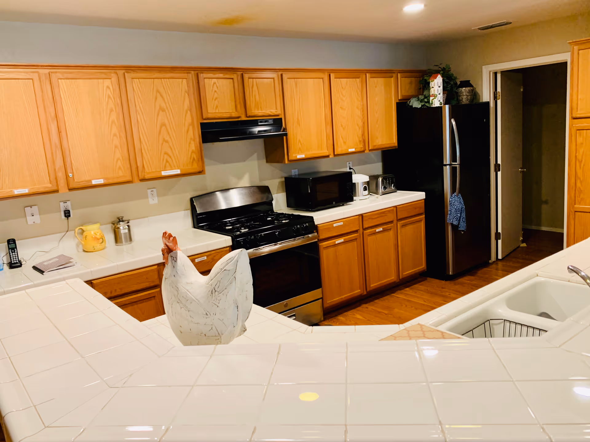 A kitchen with wooden cabinets, a stainless steel stove and refrigerator, a microwave, toaster, and coffee maker on the countertops. The foreground shows a white tiled counter with a decorative ceramic chicken. The floor is wooden, and there is a doorway leading to another room.