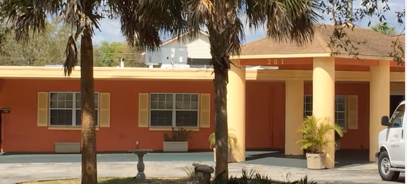 Exterior view of a single-story assisted living facility building with a red and beige color scheme, palm trees in the foreground, and a white van parked on the right side. The building has large windows with shutters and a covered entrance with the number 301 above it.