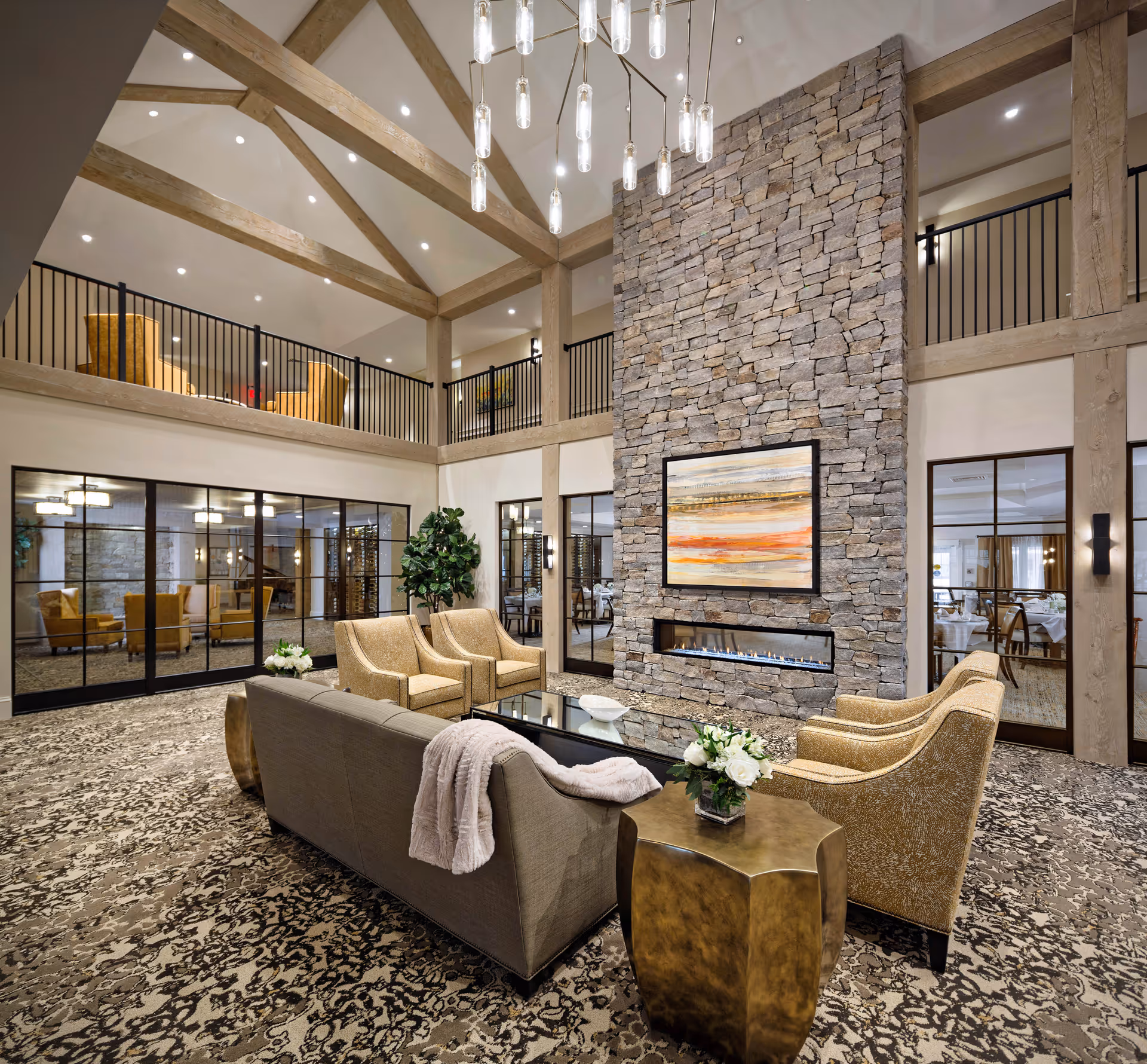 A spacious and elegant living room area with a high vaulted ceiling featuring exposed wooden beams and a modern chandelier. The room has a large stone fireplace with a horizontal gas insert and a colorful abstract painting above it. Comfortable seating includes a beige sofa with a throw blanket and four patterned armchairs arranged around a black coffee table. There are side tables with flower arrangements, patterned carpet flooring, and large windows and glass doors leading to adjacent rooms.