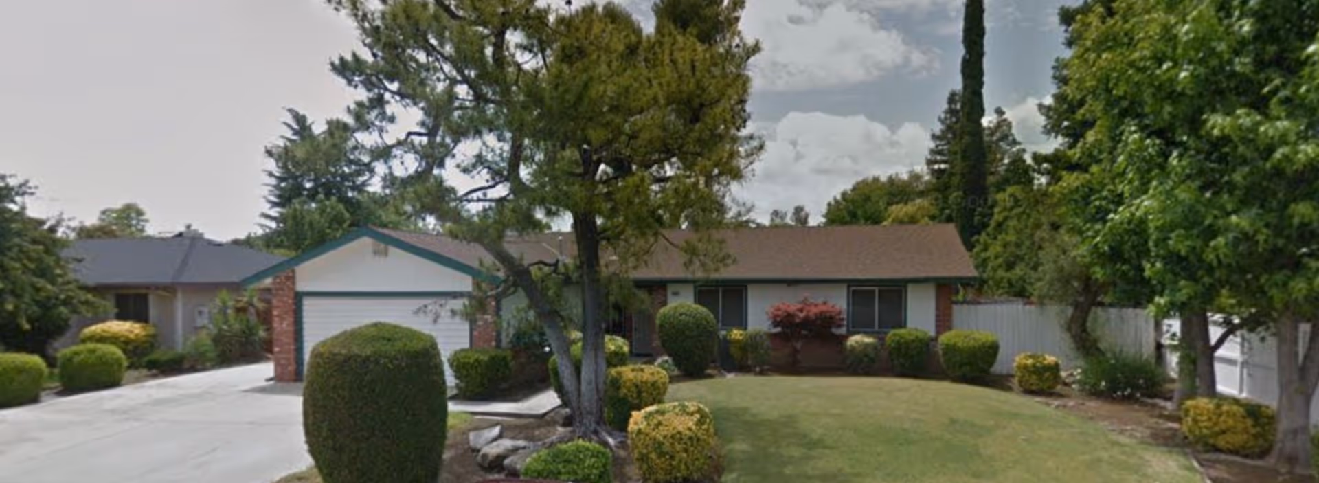 Front exterior view of a single-story residential building with a garage, surrounded by trimmed bushes, trees, and a lawn under a partly cloudy sky.