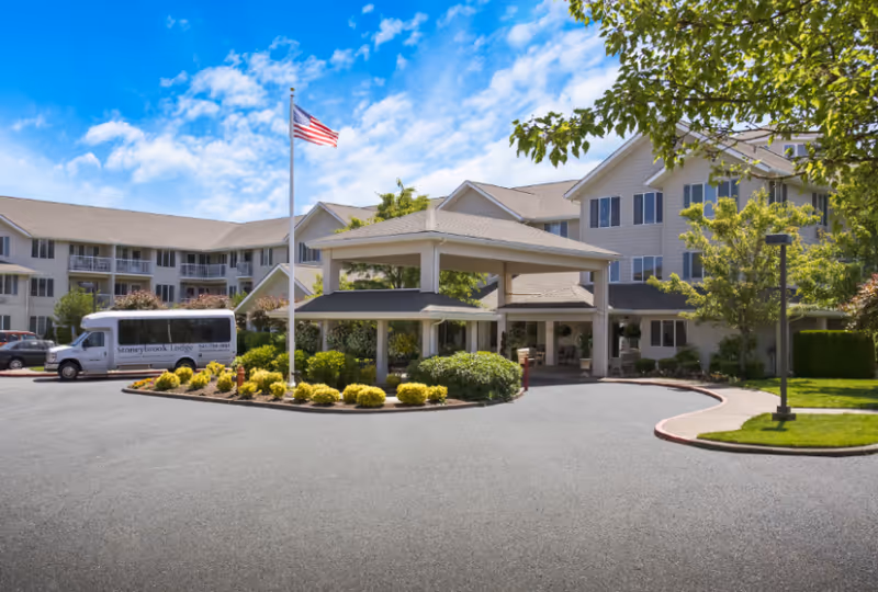 Exterior view of Stoneybrook Lodge Gracious Retirement Living facility showing a large multi-story building with beige siding, a covered entrance with a driveway, landscaped bushes and trees, an American flag on a flagpole, and a white shuttle van parked near the entrance.