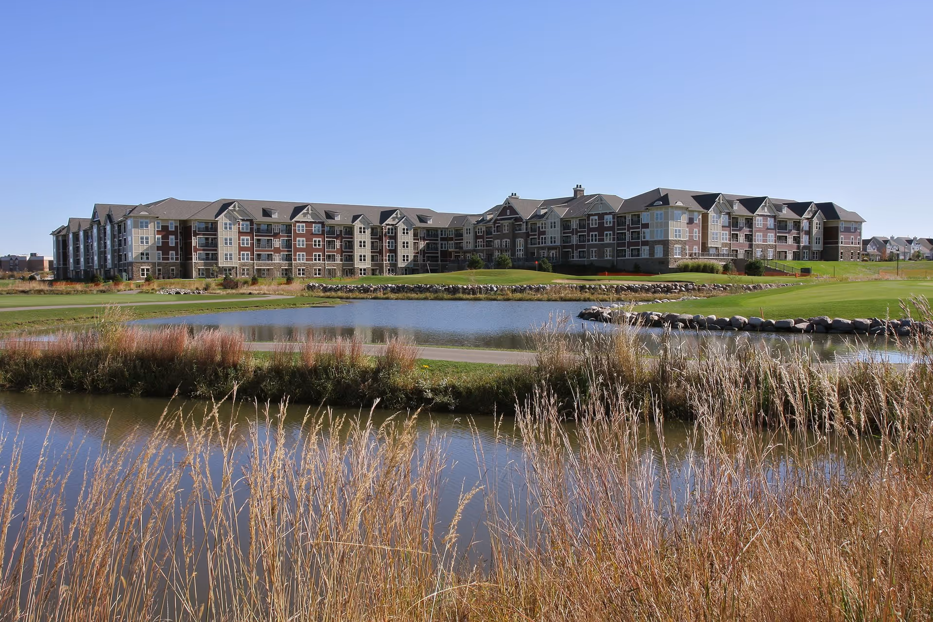 A large multi-story residential building with a mix of brick and siding exterior, situated behind a pond and grassy area with tall dry grasses in the foreground under a clear blue sky.