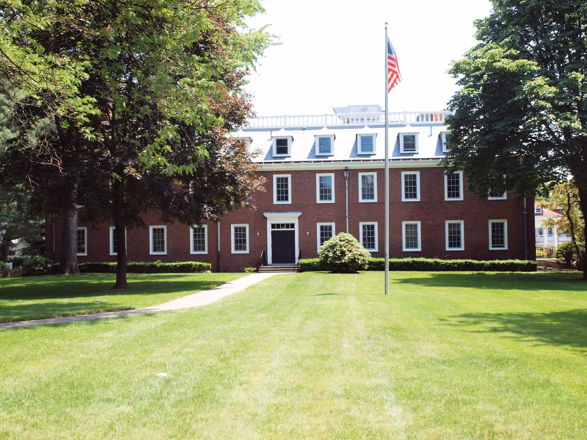 Front exterior view of a large brick building with multiple windows and a black door in the center. The building is surrounded by green grass, trees, and bushes. An American flag on a flagpole stands in front of the building.