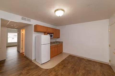 Interior view of a small kitchen area in a senior living facility with wooden cabinets, a white refrigerator, a microwave, and a sink. The kitchen has a wood floor with a linoleum section near the cabinets. Adjacent to the kitchen is a hallway leading to another room with a window.