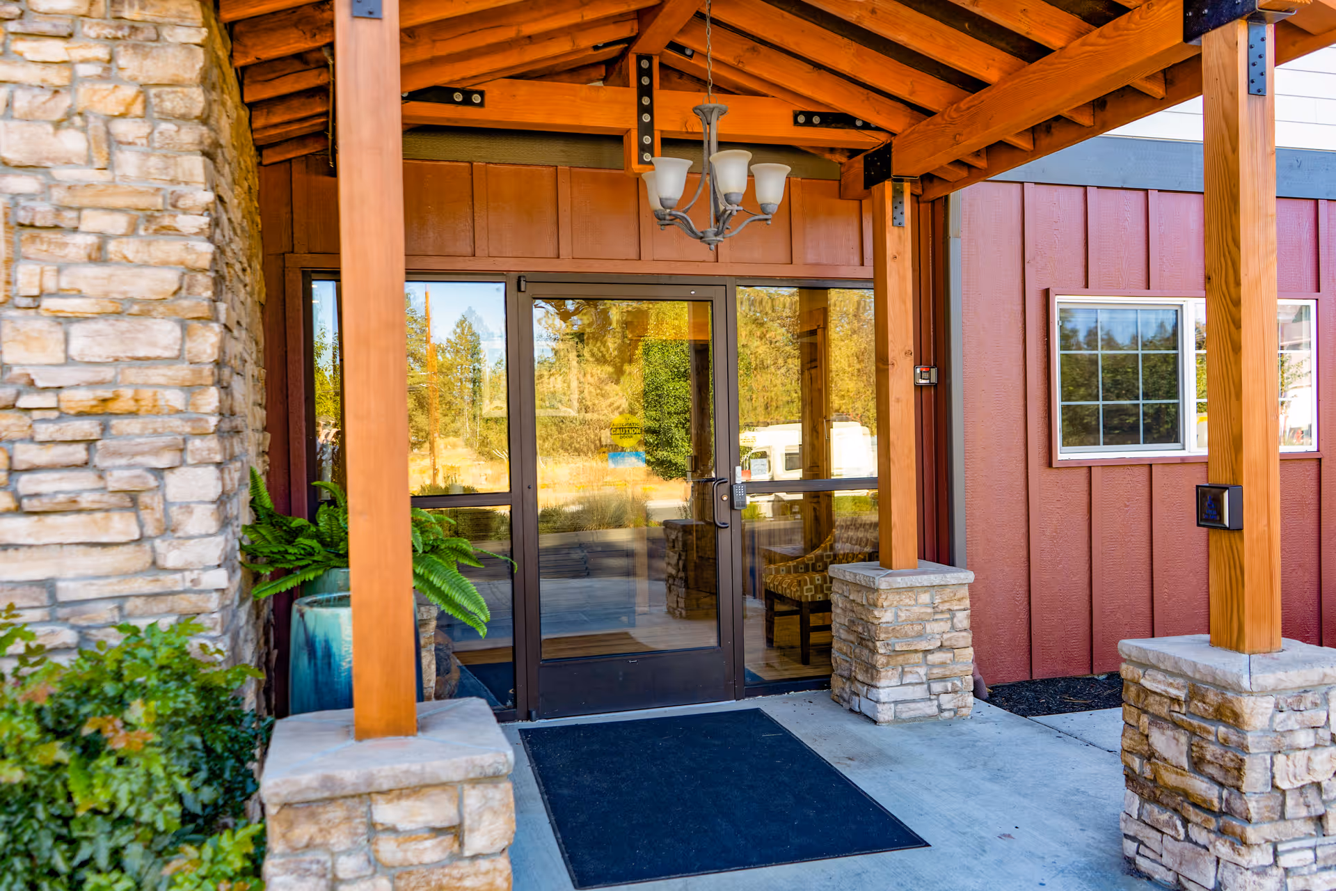 Entrance to a building with glass double doors framed by stone pillars and wooden beams. There is a black mat on the concrete floor and a chandelier hanging from the wooden ceiling. A potted plant is visible to the left of the doors, and a red exterior wall with windows is on the right.
