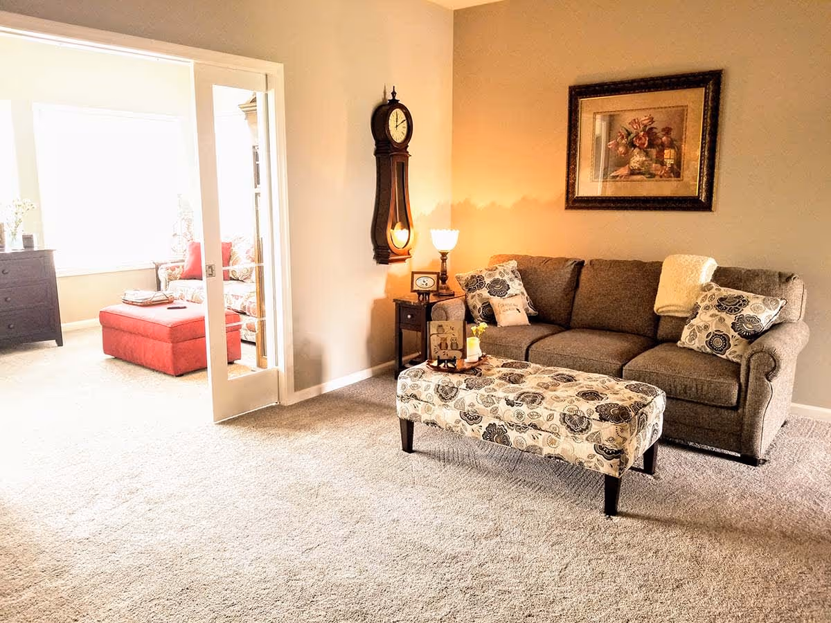 A cozy living room with a gray sofa adorned with patterned pillows and a cream-colored throw. In front of the sofa is a floral-patterned upholstered bench. A small side table with a lamp and decorative items sits next to the sofa. On the wall above the sofa is a framed floral painting. To the left, glass double doors open into a bright sunroom with a red ottoman and floral armchair. The room has beige walls and carpeted flooring.