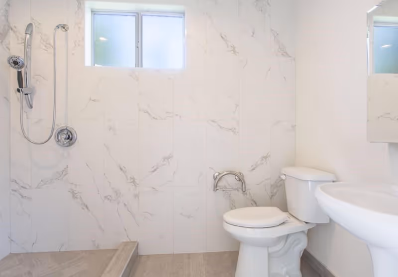 A clean bathroom with white marble-patterned tiled walls, a walk-in shower with a handheld showerhead, a white toilet with a grab bar mounted on the wall beside it, and a white pedestal sink. There is a small frosted window above the shower area.
