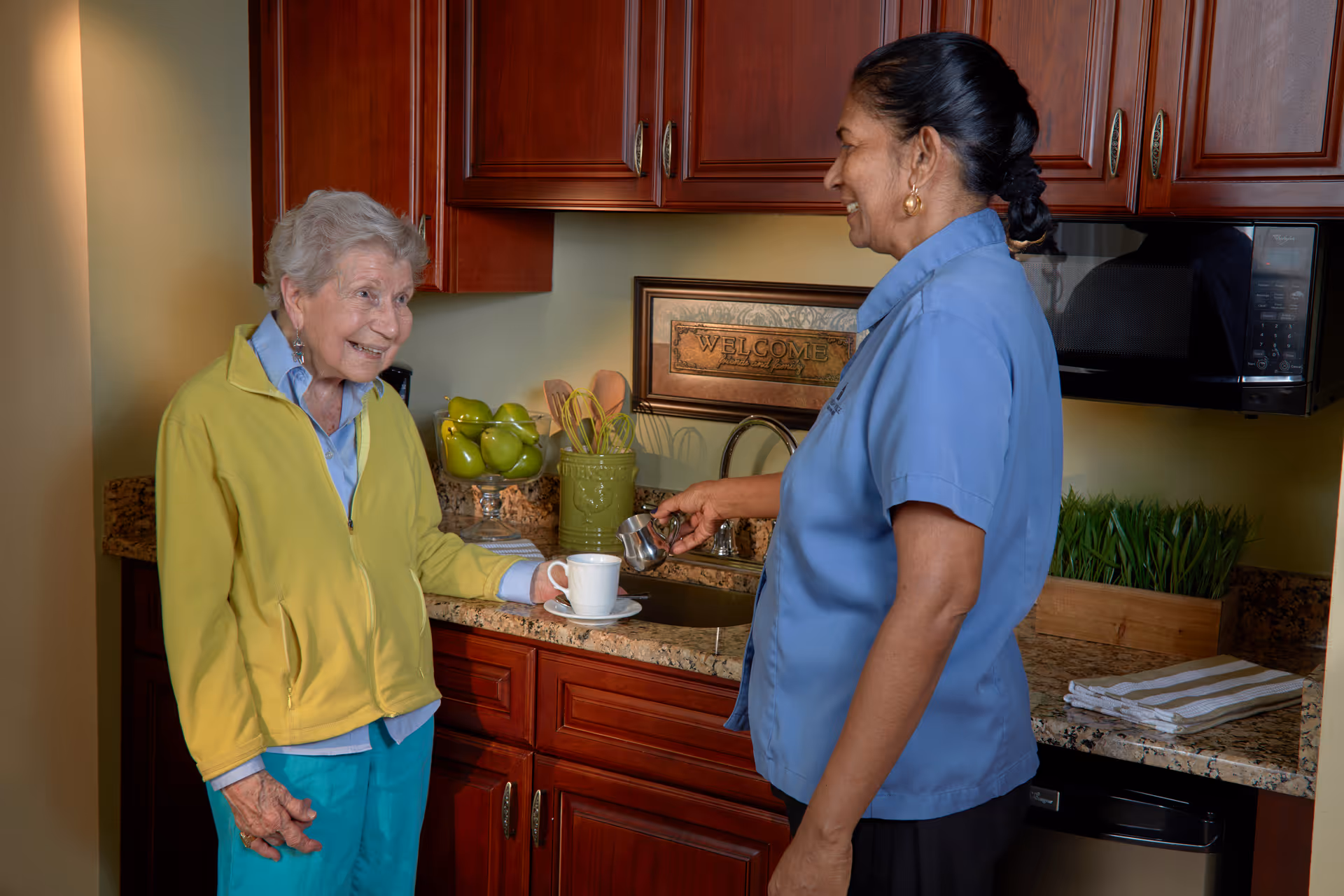 An elderly woman in a yellow jacket holding a cup and saucer smiles while a caregiver in a blue uniform pours a drink in a kitchen area with wooden cabinets and a granite countertop.