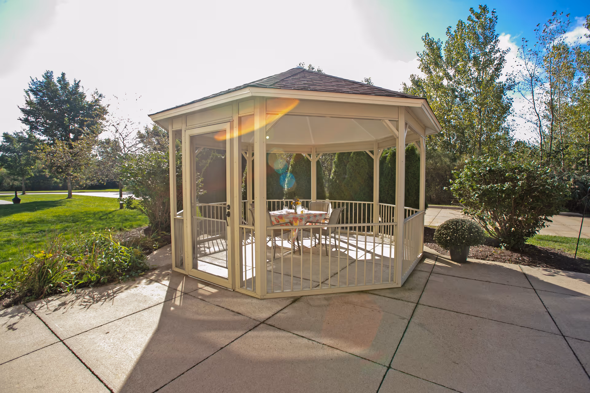 A white gazebo with a hexagonal roof situated on a concrete patio surrounded by greenery and trees. Inside the gazebo, there is a table covered with a floral tablecloth and four chairs around it. The scene is bright and sunny with lens flare visible in the image.