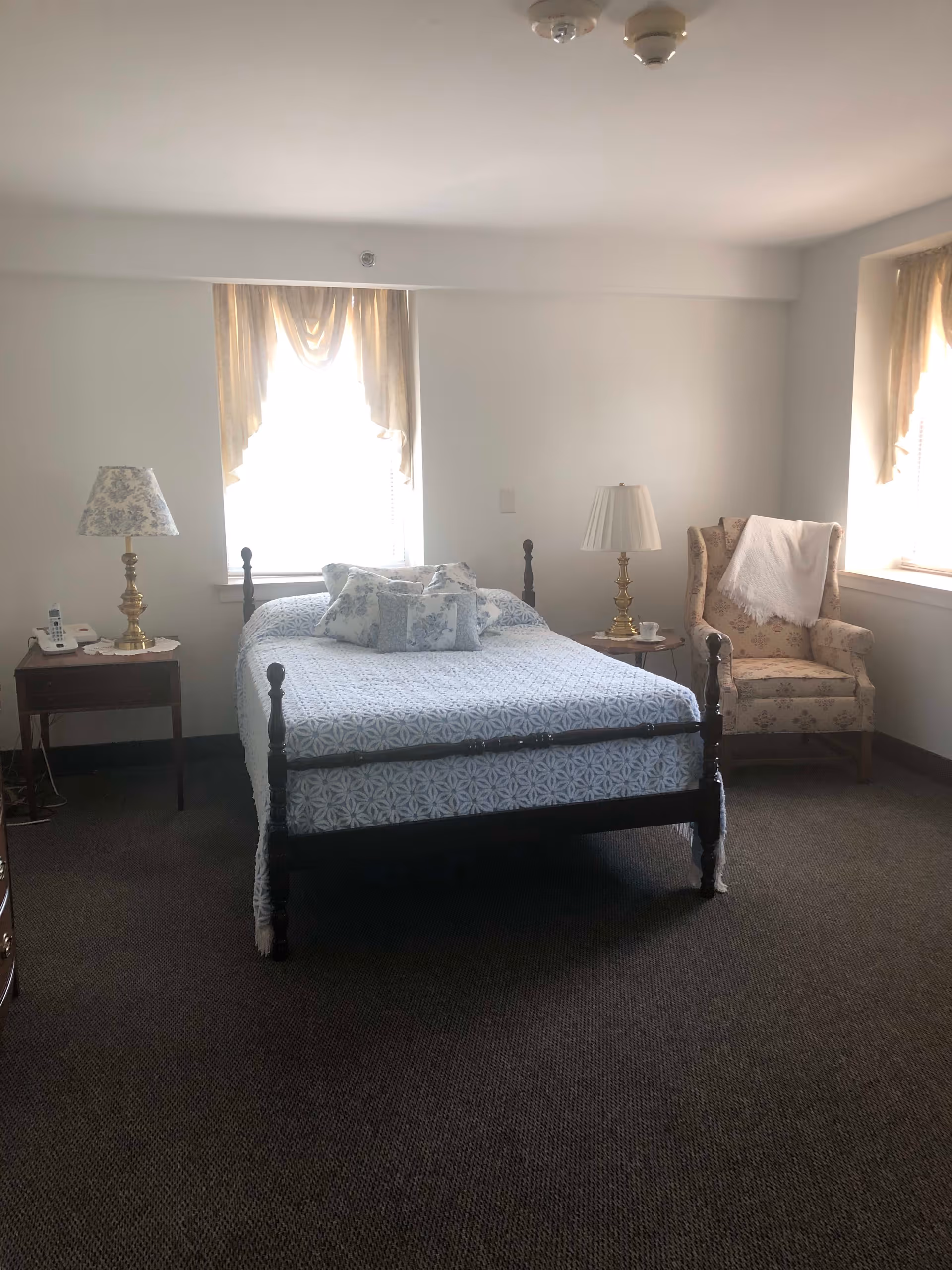 A bedroom with a wooden bed frame covered with a blue and white patterned bedspread and matching pillows. There are two windows with beige curtains letting in natural light. On the left side of the bed is a wooden nightstand with a floral lamp and a telephone. On the right side is a small round table with a white lamp and a cup. A floral upholstered armchair with a white throw blanket is positioned near the window on the right.