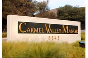 A large white stone sign with gold and black lettering that reads 'Carmel Valley Manor' with the number 8545 below it, set outdoors with grass and trees in the background.
