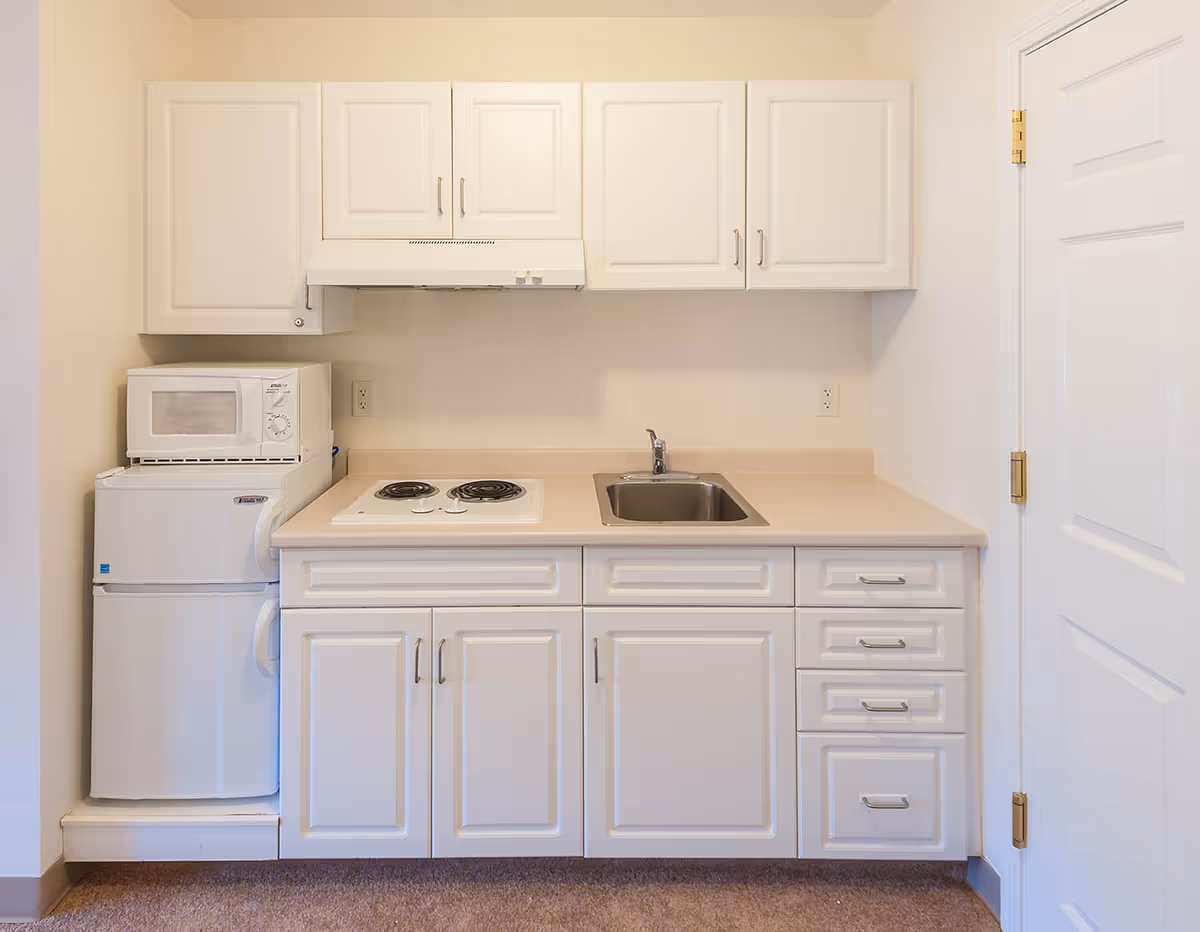 A small kitchen area with white cabinets, a countertop with a sink, a two-burner electric stove, a microwave on top of a mini refrigerator, and a white door to the right.