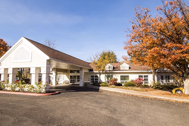 Exterior view of a single-story senior living facility building with a covered entrance, surrounded by trees with autumn foliage and a clear blue sky.
