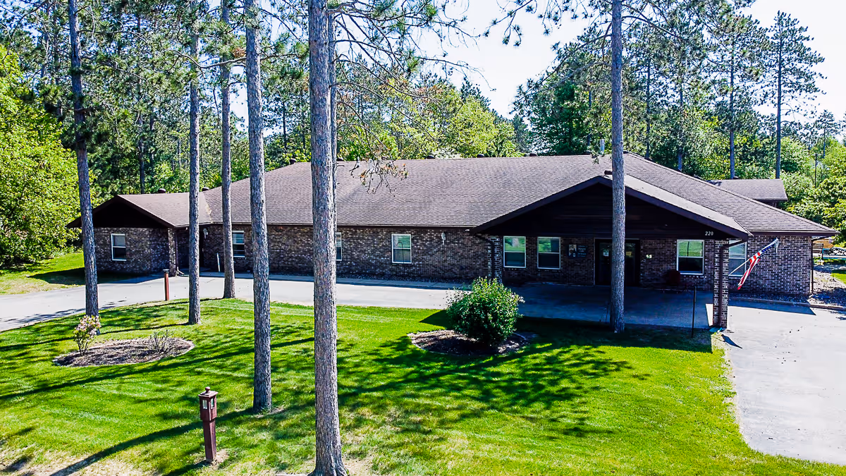 Exterior view of a single-story brick building with a brown roof surrounded by tall pine trees and green grass. There is a driveway leading to the covered entrance, and an American flag is displayed near the front door.