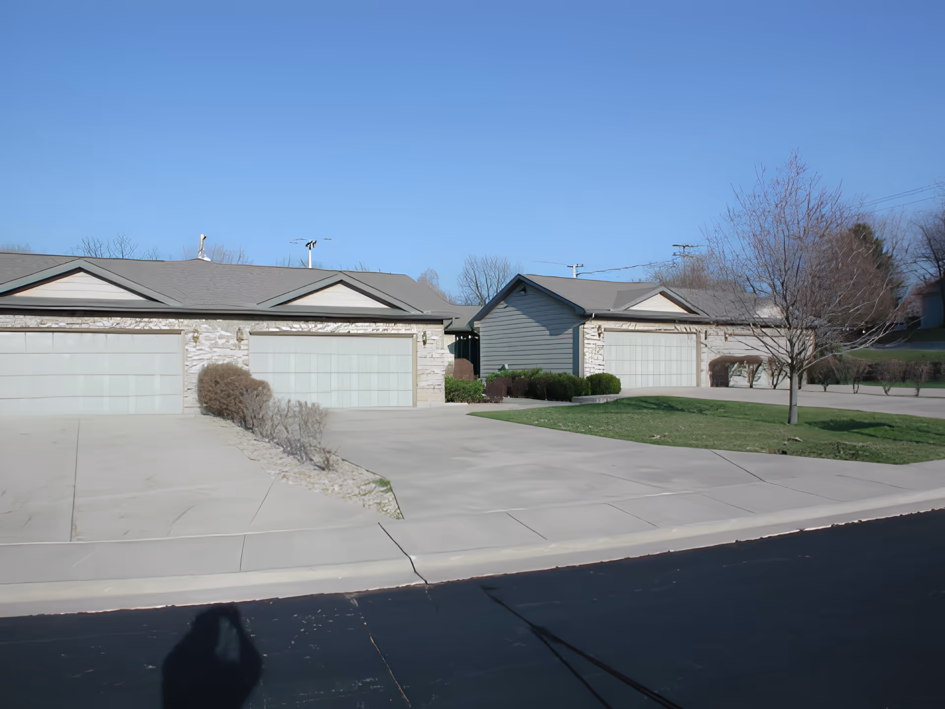 Exterior view of a senior living facility showing multiple attached garages with light-colored doors, a paved driveway, some bushes, a leafless tree, and a clear blue sky.