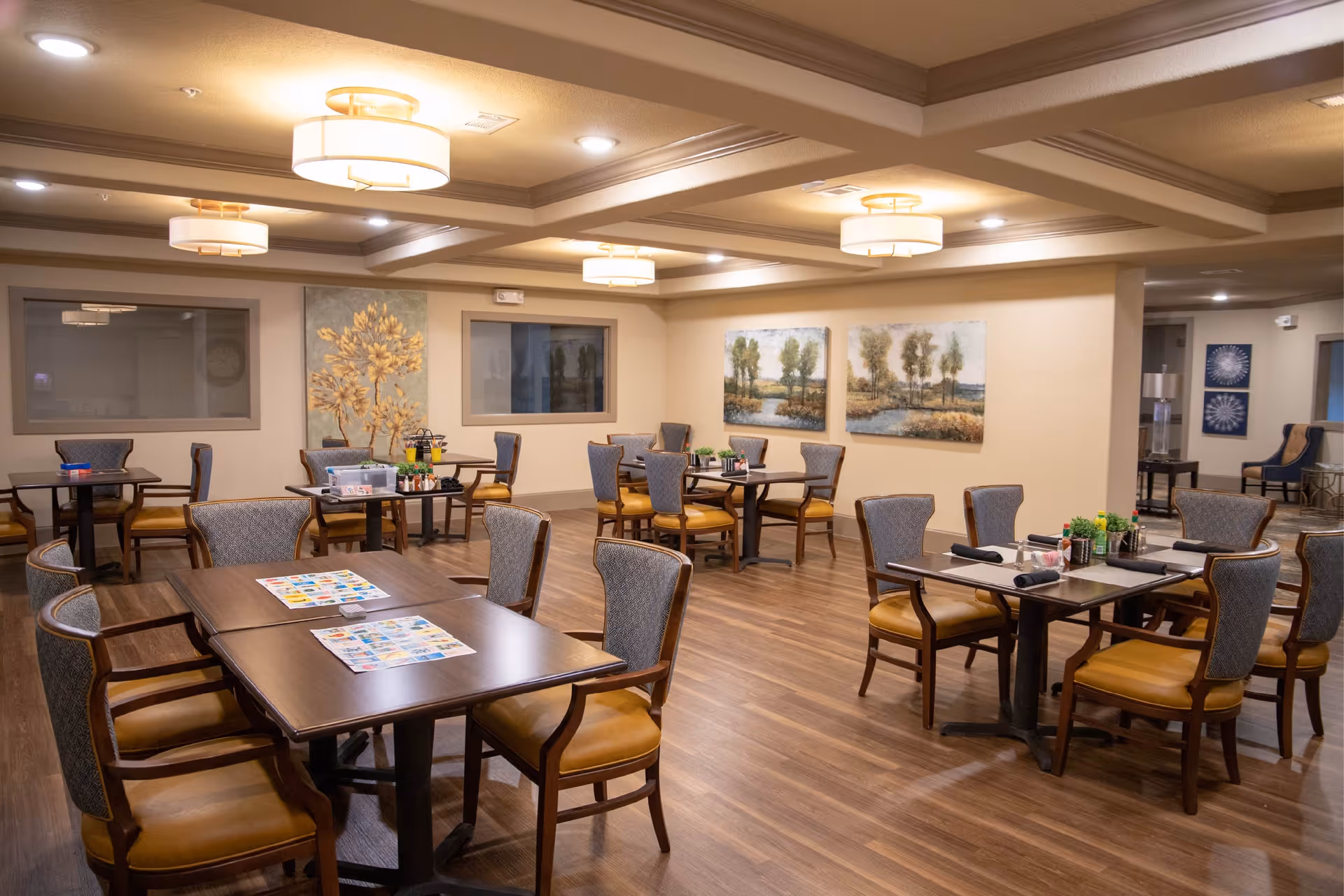 A spacious dining room with multiple wooden tables and cushioned chairs arranged neatly. The room features warm lighting with ceiling fixtures, wood flooring, and wall art depicting trees and nature scenes. The atmosphere is calm and inviting.