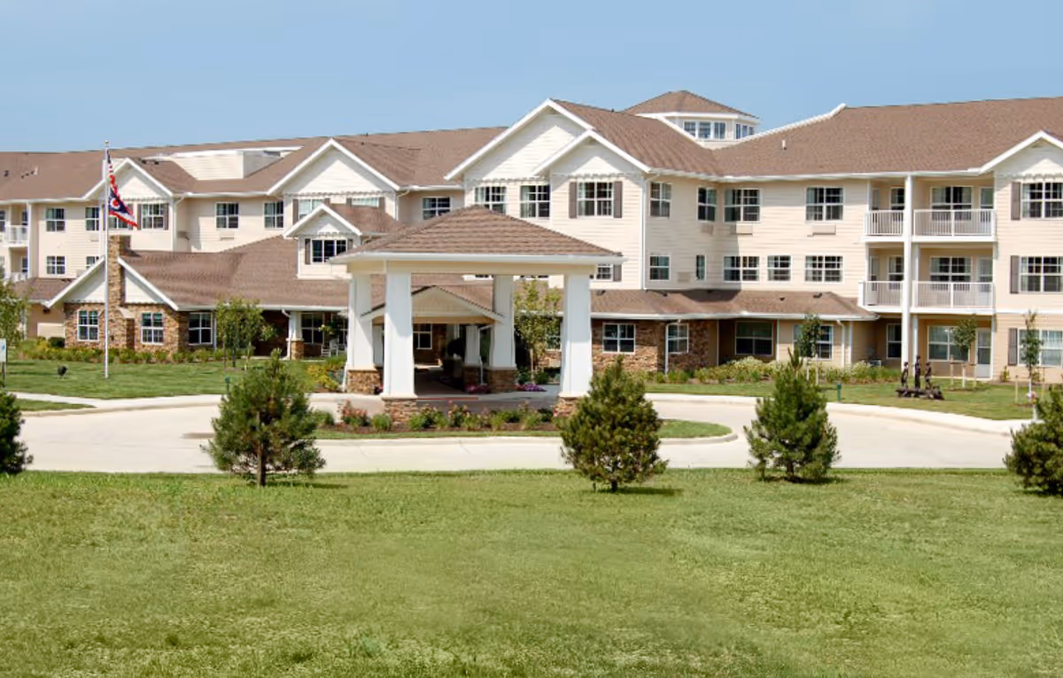 Front exterior view of Parker Place Gracious Retirement Living, a large multi-story building with beige siding and brown roofs, surrounded by green lawns and small trees, with a covered entrance and an American flag on a flagpole.