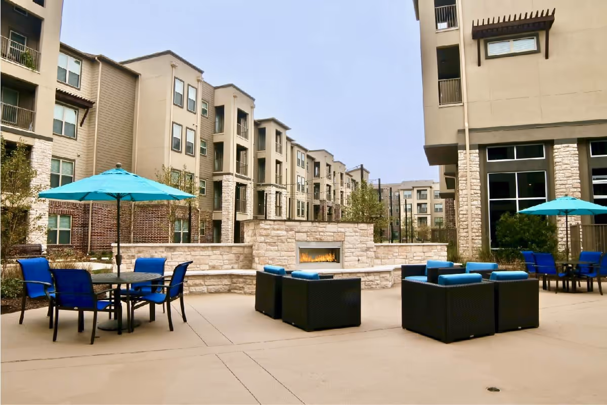 Outdoor patio area at a senior living facility with modern wicker seating featuring blue cushions, round tables with blue umbrellas, and a built-in stone fireplace. Surrounding the patio are multi-story residential buildings with balconies and large windows.