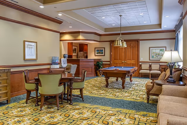 A spacious common area in a senior living facility featuring a pool table in the center, a round table with four green upholstered chairs, a beige sofa, and additional seating along the walls. The room has patterned carpet, wood paneling, framed artwork, and a ceiling with recessed lighting and a hanging light fixture above the pool table.