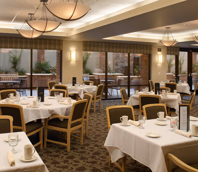 A dining room with multiple tables covered in white tablecloths, each set with cups, glasses, plates, and napkins. The room has large windows with views of an outdoor patio area with plants and brick walls. The lighting is warm with ceiling fixtures and wall sconces.