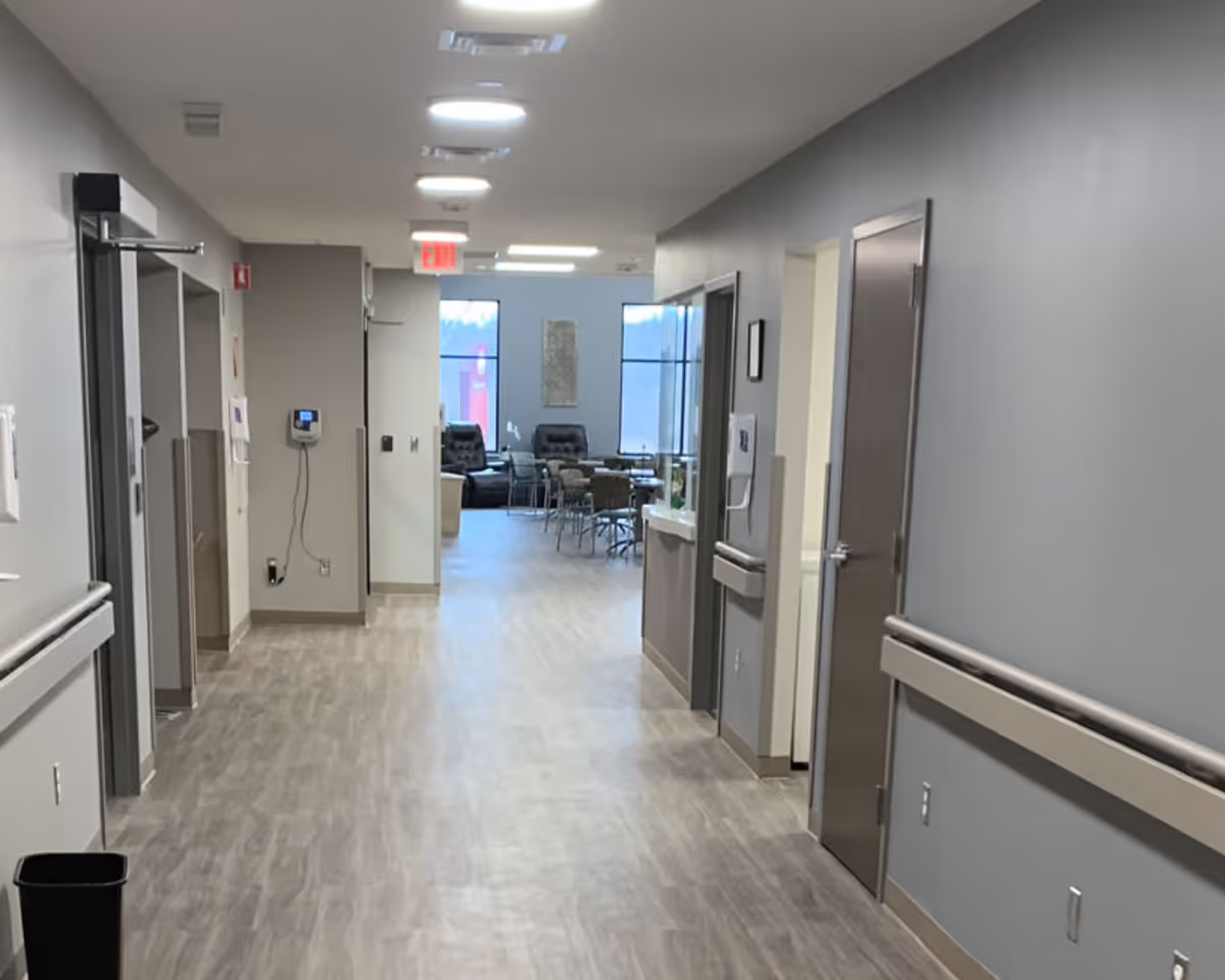A clean, well-lit hallway in a senior care facility with light wood flooring and gray walls. The hallway has handrails on both sides and several closed doors. At the end of the hallway, there is a seating area with chairs and tables near large windows letting in natural light.