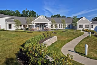 Single-story senior living facility building with a light gray exterior, white trim, and a covered entrance. The building is surrounded by a well-maintained lawn, small trees, shrubs, and a curved concrete walkway leading to the entrance under a clear blue sky.