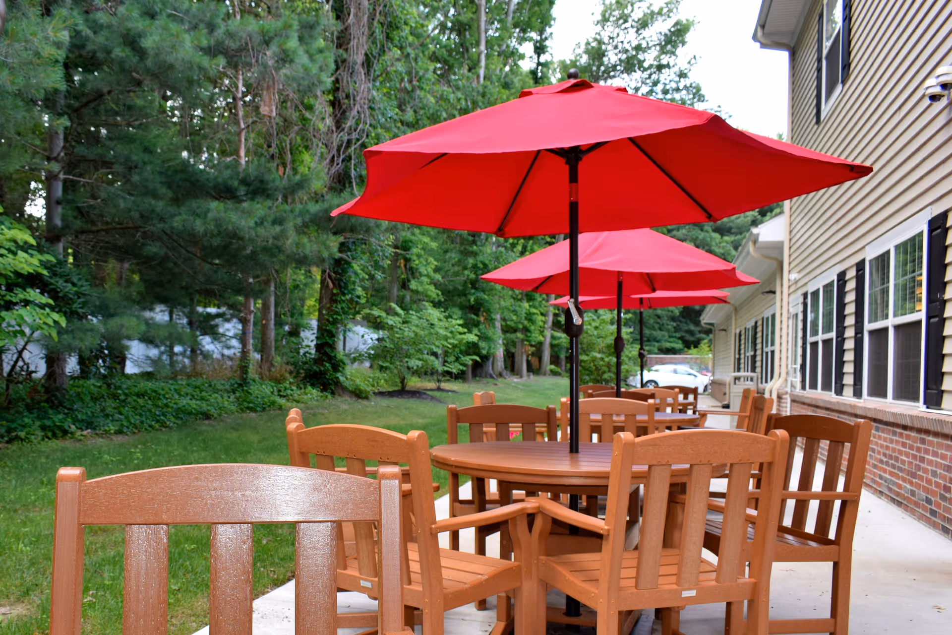 Outdoor patio area with wooden tables and chairs, each table shaded by a large red umbrella. The patio is adjacent to a building with beige siding and black shutters, surrounded by green trees and grass.